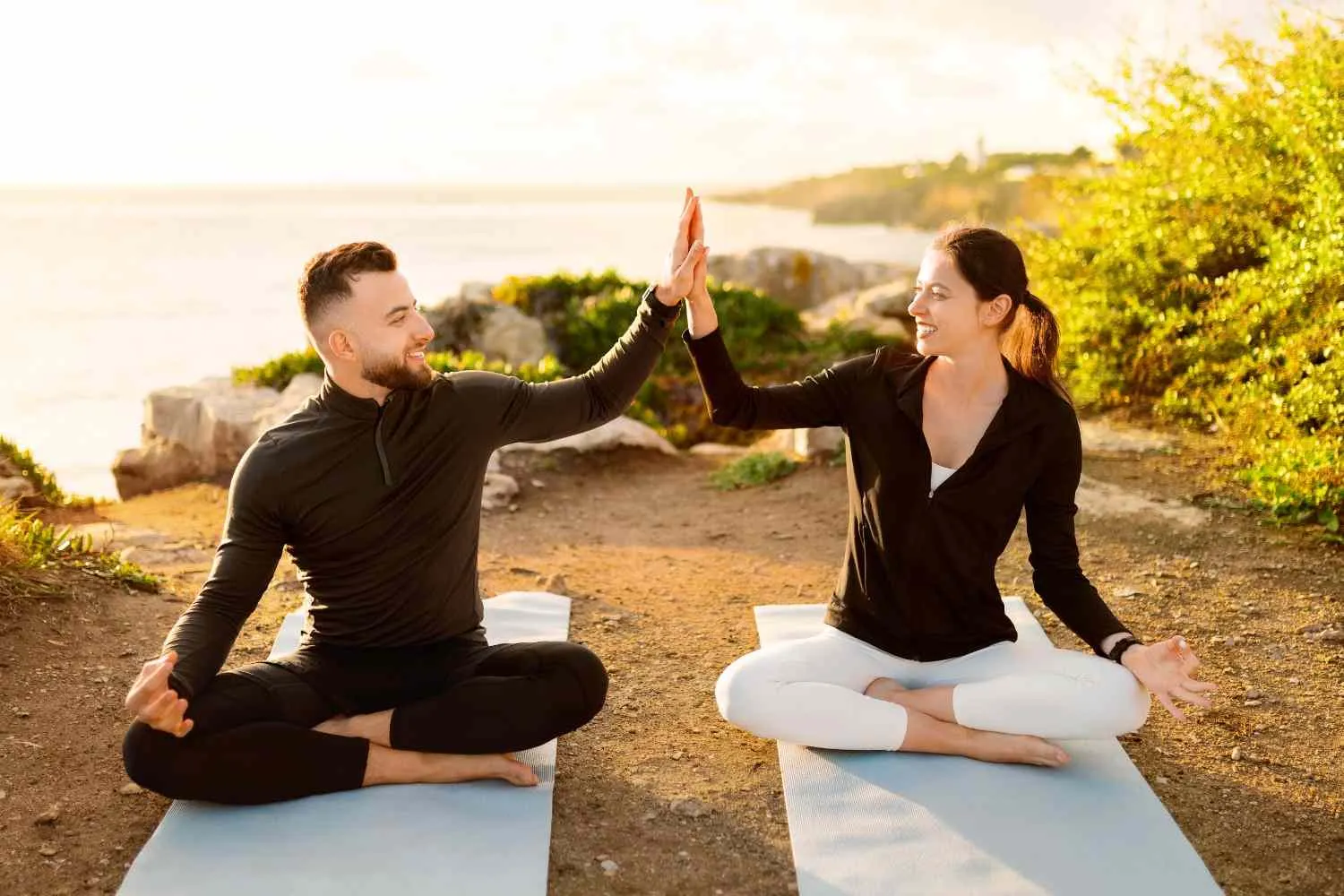 Two people on mats by the coast at sunrise touching palms above their heads
